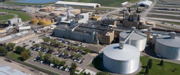 Aerial view of an industrial complex with large metal tanks on the right, a parking lot near the middle foreground, and various metal industrial buildings in the center of the image, additional tanks are on the left with a railyard and trains present, in the background is an oblong metal building and a pond