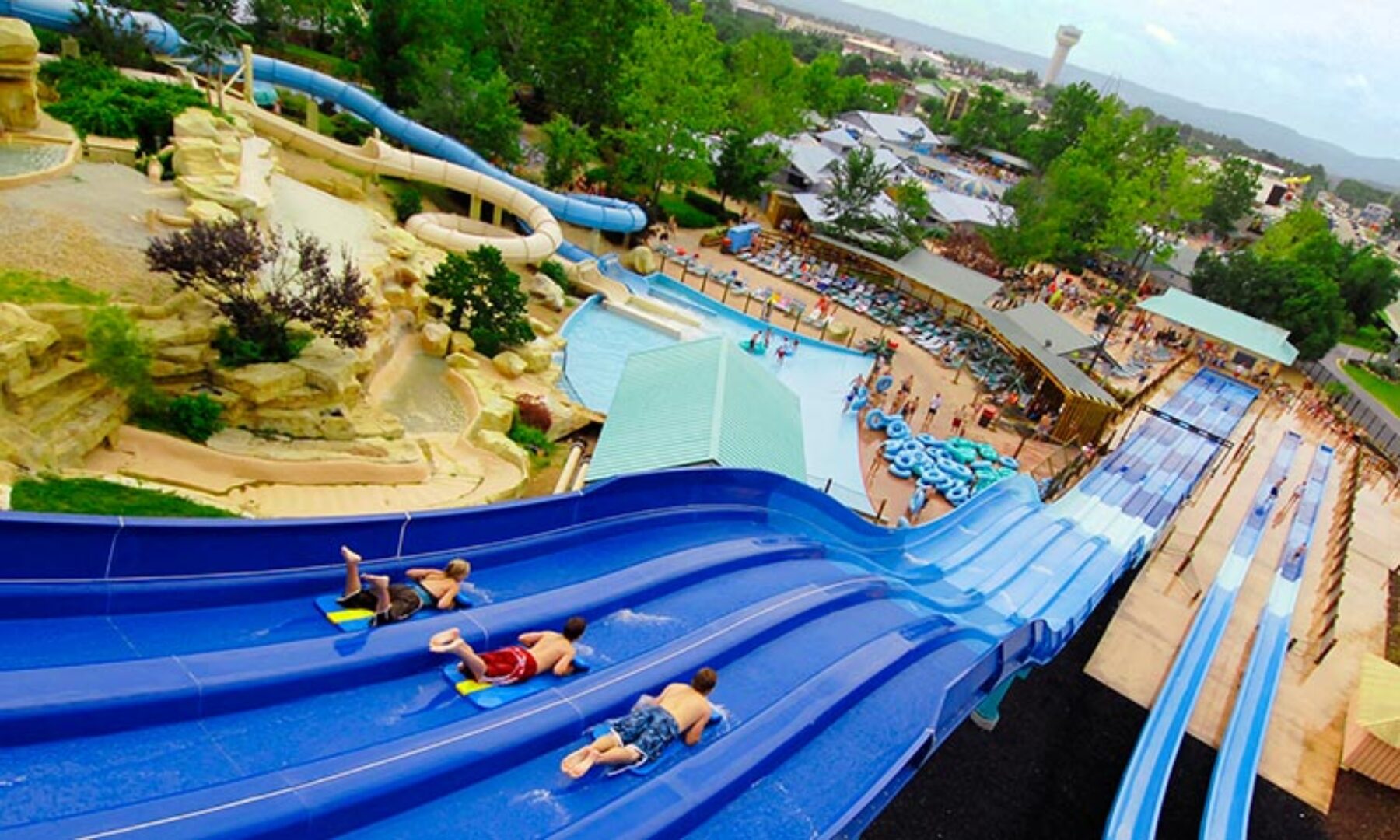 View of three lane water slide with three young people riding yellow and blue surfing mats down the slide, tube slides and swimming pools in the distance of a medium sized midwest town with a water tower in the background