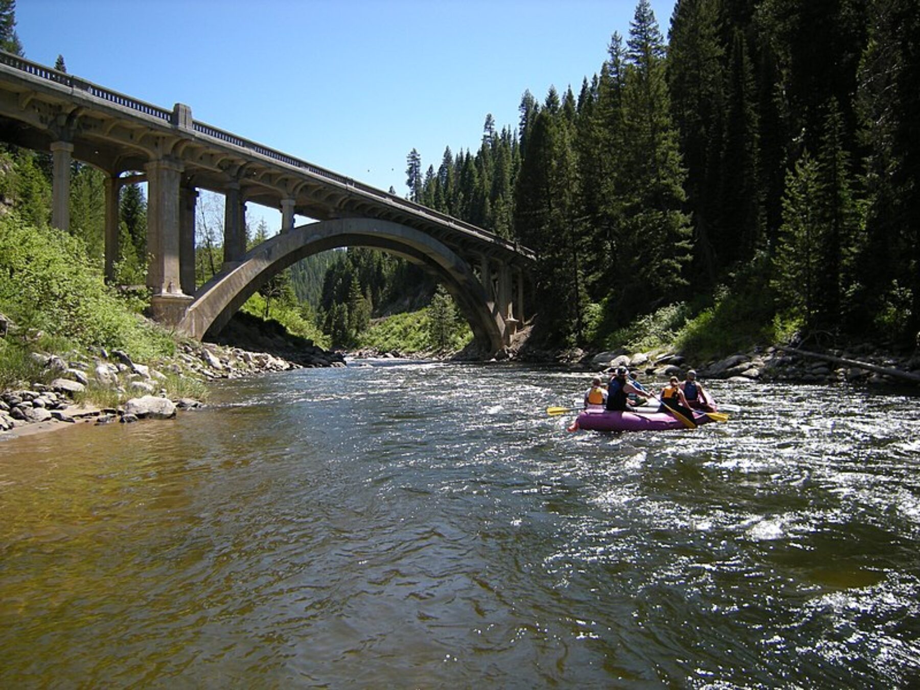 Five people of mixed genders and ages paddle a raft near Rainbow Bridge on the Payette River near North Fork Idaho with evergreen trees, shrubs, and large stones on the riverbank