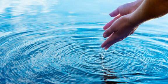 Hands pour water into a clear blue lake or pond creating ripples to accompany the blog post describing the EPA's allocation of $2.4 billion for clean water